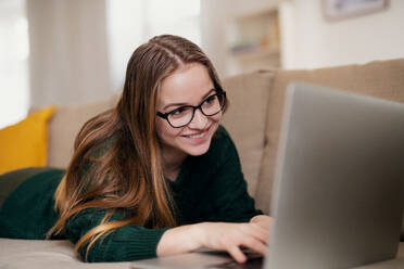 A young happy college female student lying down on sofa at home, using laptop when studying. - HPIF26310