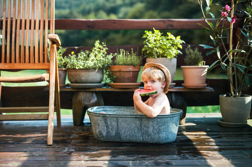 Glücklicher kleiner Junge mit Hut in der Badewanne im Garten im Sommer, isst Wassermelone. - HPIF26354