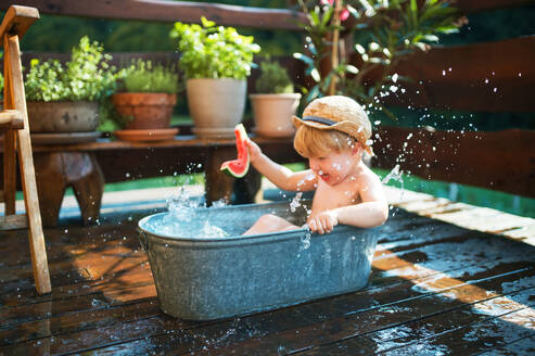 Glücklicher kleiner Junge mit Hut in der Badewanne im Garten im Sommer, isst Wassermelone. - HPIF26355