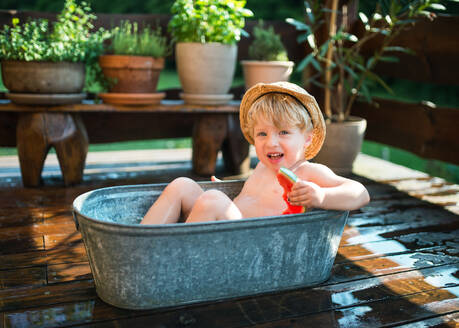 Glücklicher kleiner Junge mit Hut in der Badewanne im Garten im Sommer, isst Wassermelone. - HPIF26356