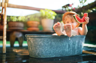 Glücklicher kleiner Junge mit Hut in der Badewanne im Garten im Sommer, isst Wassermelone. - HPIF26357