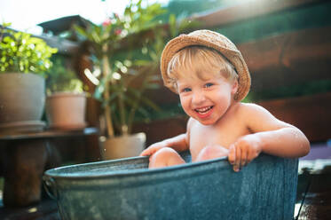 Glücklicher kleiner Junge mit Hut in der Badewanne im Garten im Sommer, der im Wasser spielt. - HPIF26365