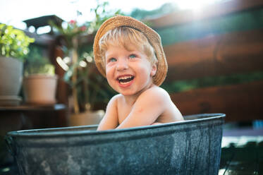 Glücklicher kleiner Junge mit Hut in der Badewanne im Garten im Sommer, der im Wasser spielt. - HPIF26366
