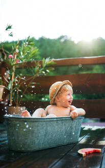 Glücklicher kleiner Junge mit Hut in der Badewanne im Garten im Sommer, isst Wassermelone. - HPIF26370