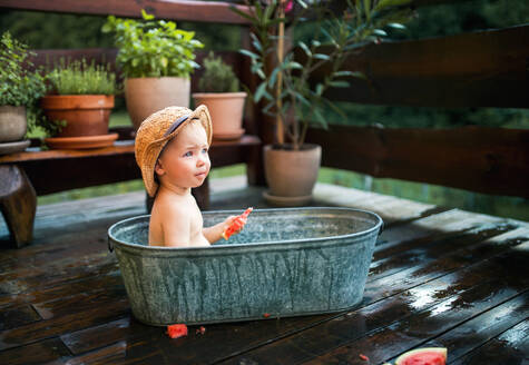 Glücklicher kleiner Junge mit Hut in der Badewanne im Garten im Sommer, isst Wassermelone. - HPIF26372