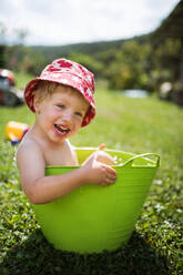 Kleiner Junge mit Hut sitzt im Plastikeimer im Garten im Sommer und spielt im Wasser. - HPIF26384