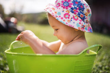 Kleines Mädchen mit Hut sitzt in Plastikeimer draußen im Garten im Sommer, spielt im Wasser. - HPIF26385