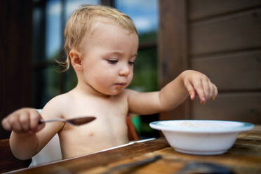 Niedliches kleines Mädchen oben ohne, das an einem Tisch auf einer Sommerterrasse steht und eine Suppe isst. - HPIF26386