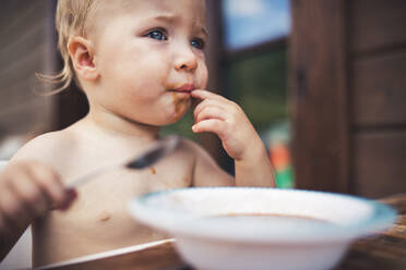 Cute topless small girl standing by a table on a patio in summer, eating soup. - HPIF26388