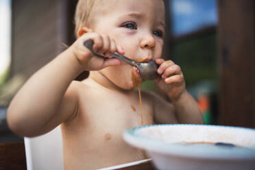 Niedliches kleines Mädchen oben ohne, das an einem Tisch auf einer Sommerterrasse steht und eine Suppe isst. - HPIF26389