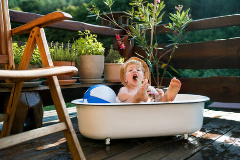 Glücklicher kleiner Junge mit Hut in der Badewanne im Garten im Sommer, der im Wasser spielt. - HPIF26393
