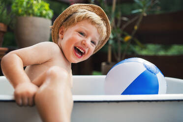 Glücklicher kleiner Junge mit Hut in der Badewanne im Garten im Sommer, der im Wasser spielt. - HPIF26395