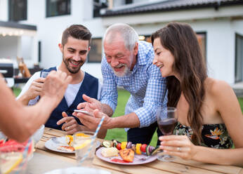 Eine Gruppe von Menschen genießt ein Barbecue im Garten einer Familie und hält den Moment mit einem Selfie auf ihrem Smartphone fest - HPIF26493