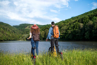 Rückansicht eines jungen Touristenpaares, das mit einem Elektroroller in der Natur unterwegs ist und am See steht. - HPIF26633