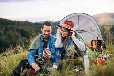 Junges Touristenpaar mit Zeltunterkunft, das abends in der Natur sitzt und sein Smartphone benutzt. - HPIF26668