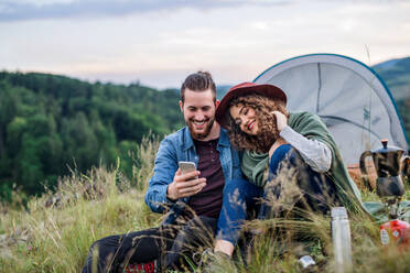 Junges Touristenpaar mit Zeltunterkunft, das abends in der Natur sitzt und sein Smartphone benutzt. - HPIF26670