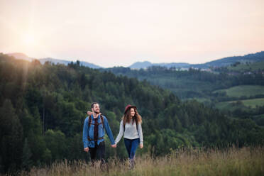 Young tourist couple travellers with backpacks hiking in nature at dusk, holding hands. - HPIF26688