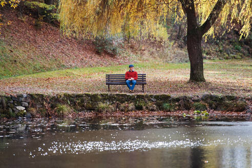 Ein einsamer älterer Mann sitzt auf einer Bank am See in der Natur und schaut in die Kamera. - HPIF26694