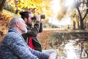 Älterer Vater und sein Sohn mit Fernglas sitzen auf einer Bank in der Natur und unterhalten sich. - HPIF26707