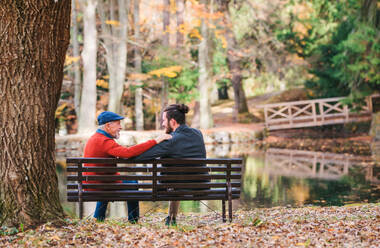 Rückansicht eines älteren Vaters und seines Sohnes, die auf einer Bank am See in der Natur sitzen und sich unterhalten. - HPIF26738