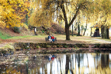 Älterer Vater und sein kleiner Sohn sitzen auf einer Bank am See in der Natur und unterhalten sich. - HPIF26739