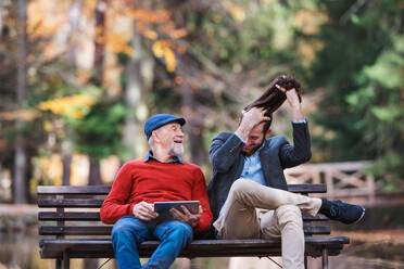 Ein älterer Vater und sein Sohn sitzen auf einer Bank in der Natur und benutzen ein Tablet. - HPIF26743