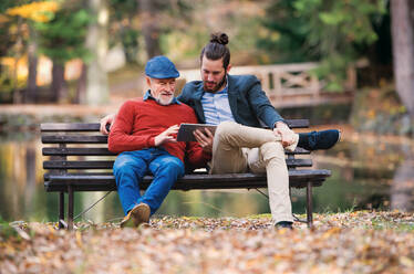 Ein älterer Vater und sein Sohn sitzen auf einer Bank in der Natur und benutzen ein Tablet. - HPIF26744