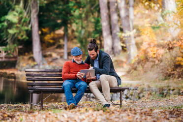 Ein älterer Vater und sein Sohn sitzen auf einer Bank in der Natur und benutzen ein Tablet. - HPIF26745