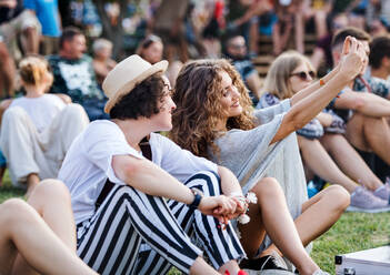 Eine Gruppe glücklicher junger Freunde, die bei einem Sommerfest auf dem Boden sitzen und einen Moment mit einem Selfie festhalten - HPIF26827
