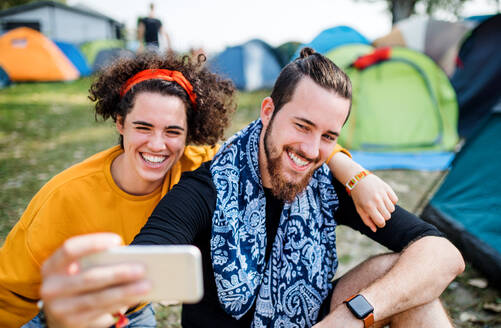 Front view of young couple at summer festival or camping holiday, taking selfie with smartphone. - HPIF26858