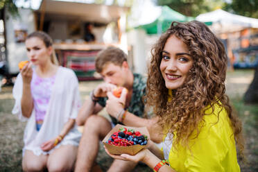 Schönes junges Mädchen mit Freunden sitzt auf dem Boden beim Sommerfest und isst Obst. - HPIF26867