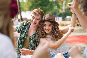 Group of cheerful young friends sitting on ground at summer festival, dancing. - HPIF26894