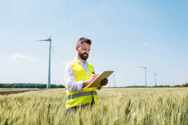 Ein Ingenieur oder Techniker mit Klemmbrett steht auf einem Feld in einem Windpark und macht sich Notizen. - HPIF26976