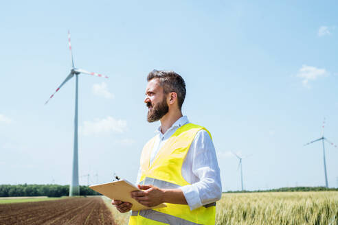 Ein Ingenieur oder Techniker mit Klemmbrett steht auf einem Feld in einem Windpark und macht sich Notizen. - HPIF26985