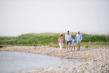 Eine Mehrgenerationenfamilie, die im Urlaub am See spazieren geht und sich an den Händen hält. - HPIF27020