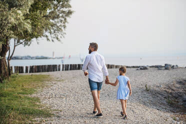 Rückansicht eines reifen Vaters, der mit seiner kleinen Tochter im Urlaub am See spazieren geht und die Hände hält. - HPIF27050
