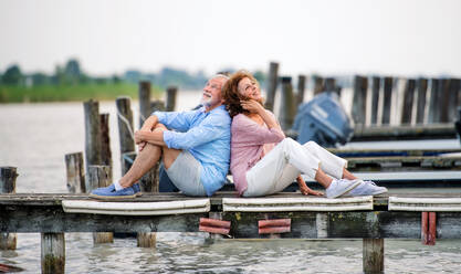Fröhliches Seniorenpaar im Urlaub, das Rücken an Rücken am See sitzt. - HPIF27075