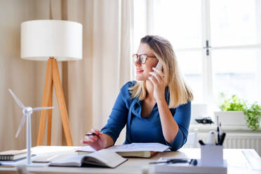 Eine junge Frau Architektin mit Smartphone sitzen am Schreibtisch drinnen im Büro zu Hause, arbeiten. - HPIF27093