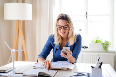 A young woman architect with smartphone sitting at the desk indoors in home office, working. - HPIF27094