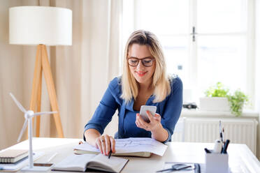 Eine junge Frau Architektin mit Smartphone sitzen am Schreibtisch drinnen im Büro zu Hause, arbeiten. - HPIF27094