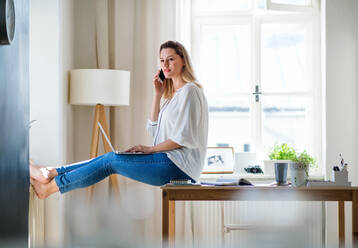 A young woman with smartphone and laptop indoors in home office, working. - HPIF27103