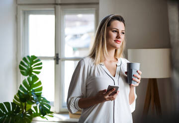 Young woman with smartphone standing indoors in home office, holding drink. - HPIF27105