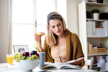 Young woman sitting at the table indoors at home, reading a book and eating. - HPIF27111