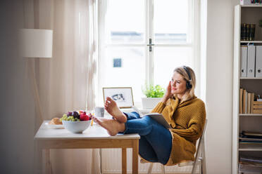 Glückliche junge Frau mit Kopfhörern, die zu Hause am Tisch sitzt und ein Buch liest. - HPIF27119