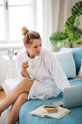 Eine junge Frau mit Kaffee auf dem Bett sitzend zu Hause am Morgen, mit Laptop. - HPIF27129