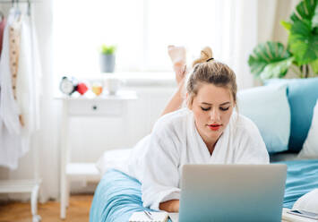 A young woman lying on bed indoors at home in the morning, using laptop. - HPIF27131