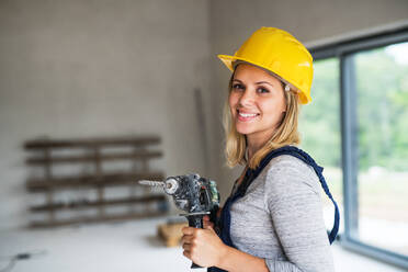 Portrait of a female worker holding an electirc drill on the construction site. Close-up of a beautiful young woman with a yellow helmet. Copy space. - HPIF27235