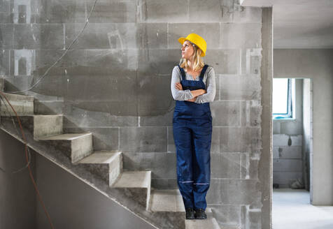 Junge Arbeiterin mit gelbem Helm, die mit verschränkten Armen auf der Treppe einer Baustelle steht, Raum kopieren. - HPIF27244