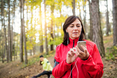 Zwei weibliche Läuferinnen mit Smartphone und Kopfhörer stehen draußen im Wald in der herbstlichen Natur und ruhen sich aus. - HPIF27268