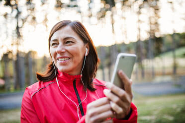 Eine alte Läuferin mit Kopfhörern und Smartphone im Freien in der herbstlichen Natur, die Musik hört. - HPIF27300